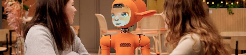 Two women talk to an orange robot at a table.