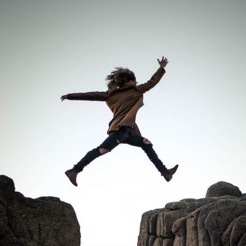 person jumping on big rock under gray and white sky during daytime